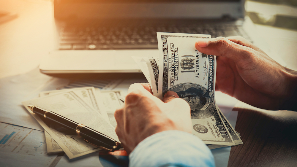 Hands counting $100 dollar bills with laptop in background