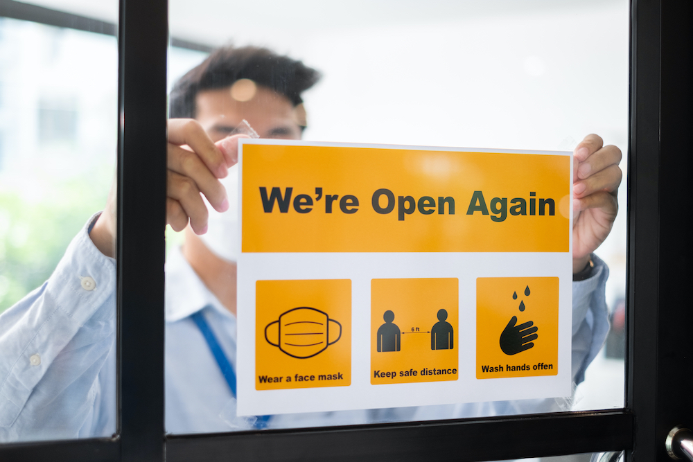 Man hanging sign in widow that says 'We are Open Again' with informational icons below for COVID safety