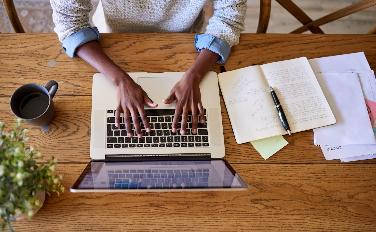 Overhead view of hands typing on a laptop keyboard with notebook to side