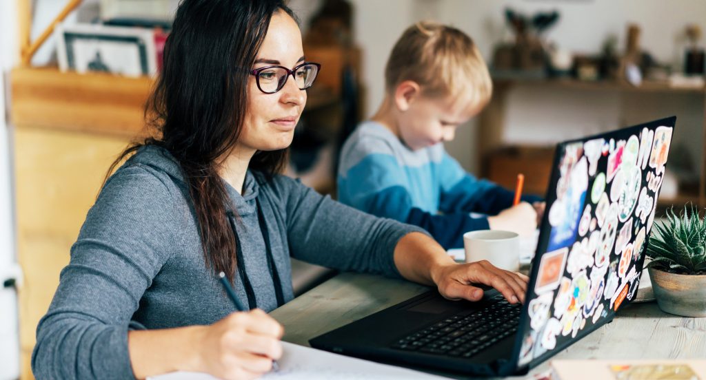 Woman virtual learning, sitting at table with laptop her child sits next to her writing or drawing.