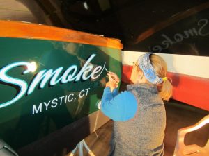 Sarah Brandon hand lettering the name Smoke on the back of a boat