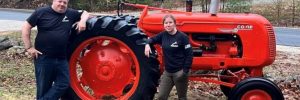 Two white people standing in front of a red tractor in rural Maine, leaves and grass on ground with pine trees in the background.