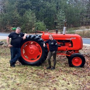 Two white people standing in front of a red tractor in rural Maine, leaves and grass on ground with pine trees in the background.