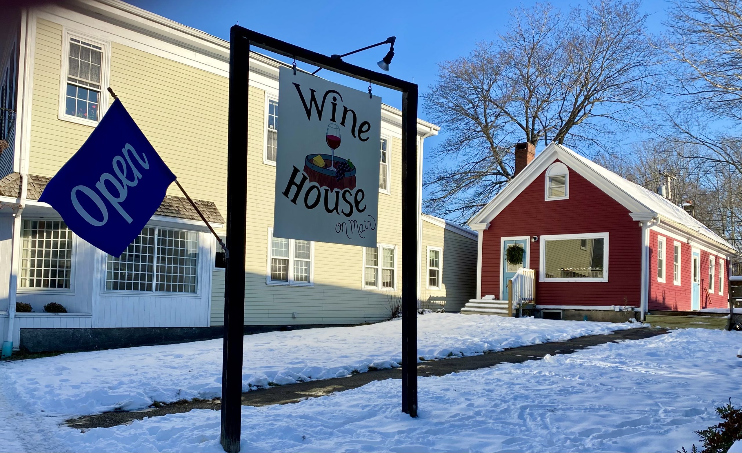 External photo of Wine House on Main's red building and large sign with logo and open flag. 