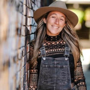 Brinn Flagg, a white women with blonde hair leans on a wooden wall. Wearing a tan wide brimmed hat, denim overalls, and a dark patterned long sleeve shirt.