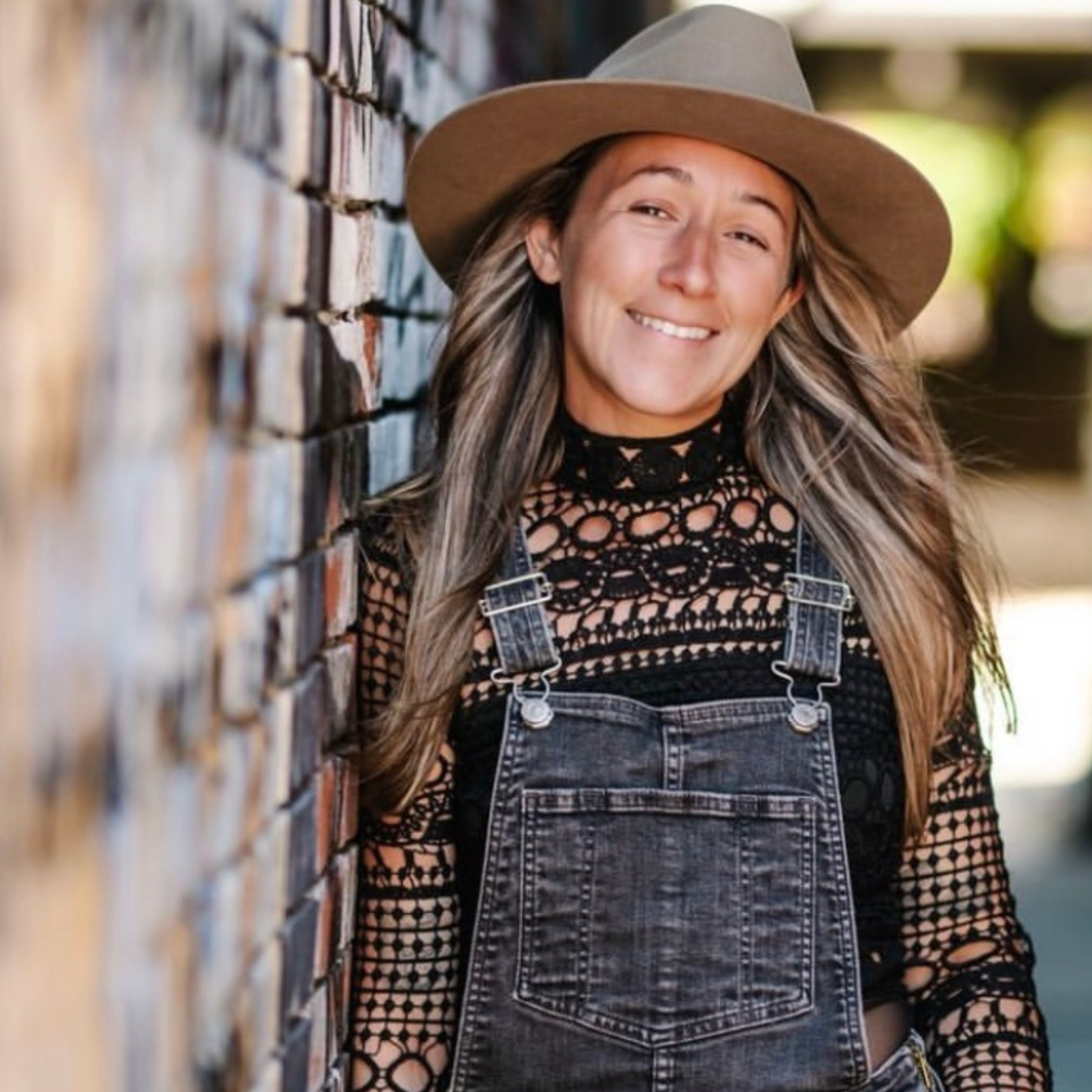 Brinn Flagg, a white women with blonde hair leans on a wooden wall.  Wearing a tan wide brimmed hat, denim overalls, and a dark patterned long sleeve shirt. 