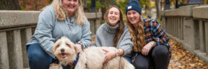 Three white women squat on a wooded trail with a white and tan dog, all 4 are smiling.