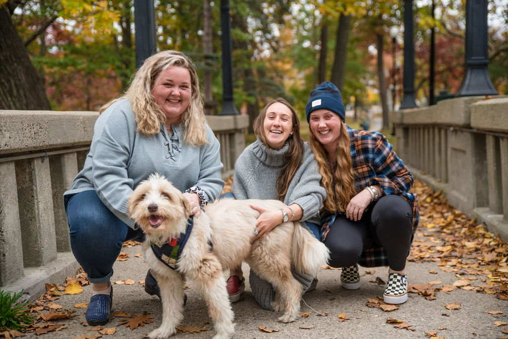 Three white women squat on a wooded trail with a white and tan dog, all 4 are smiling. 