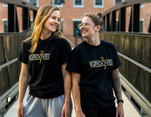 Groove 207 owners, two women smiling at each other wearing shirts with Groove 207 Logo, white and gold type on black background
