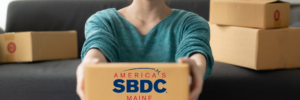 Women of Asian decent sits crisscrossapplesauce on the floor in a loose teal long sleeve shirt and blue jeans. She is surrounded by boxes, and is holding one box out in front of her. It has the Maine SBDC logo on the front of the box. It is a white room with natural light on a dark grey couch and a few plants.