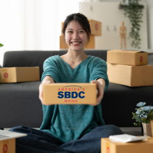 Women of Asian decent sits crisscrossapplesauce on the floor in a loose teal long sleeve shirt and blue jeans. She is surrounded by boxes, and is holding one box out in front of her. It has the Maine SBDC logo on the front of the box. It is a white room with natural light on a dark grey couch and a few plants.