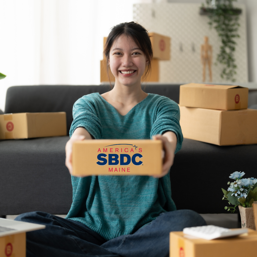 Women of Asian decent sits crisscrossapplesauce on the floor in a loose teal long sleeve shirt and blue jeans. She is surrounded by boxes, and is holding one box out in front of her. It has the Maine SBDC logo on the front of the box. It is a white room with natural light on a dark grey couch and a few plants. 