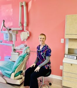 a smiling white women in colorful scrubs sits in a pink room with a teal dentist chair