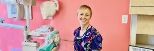 a smiling white women in colorful scrubs sits in a pink room with a teal dentist chair