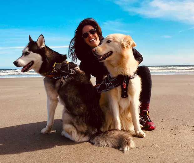 Stephanie Smith of Sacred Paws smiles while squatting on a sunny sandy beach with two dogs. 