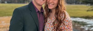 Noah and Ashley Sixberry, a young couple stand close to each other outside in front of a Maine background.