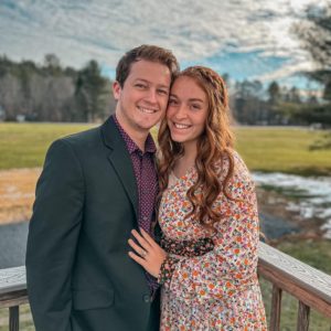 Noah and Ashley Sixberry, a young couple stand close to each other outside in front of a Maine background.