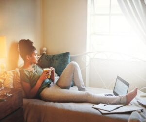Woman lounging on couch with tea in hand, gazing reflectively out the window. Laptop nearby.