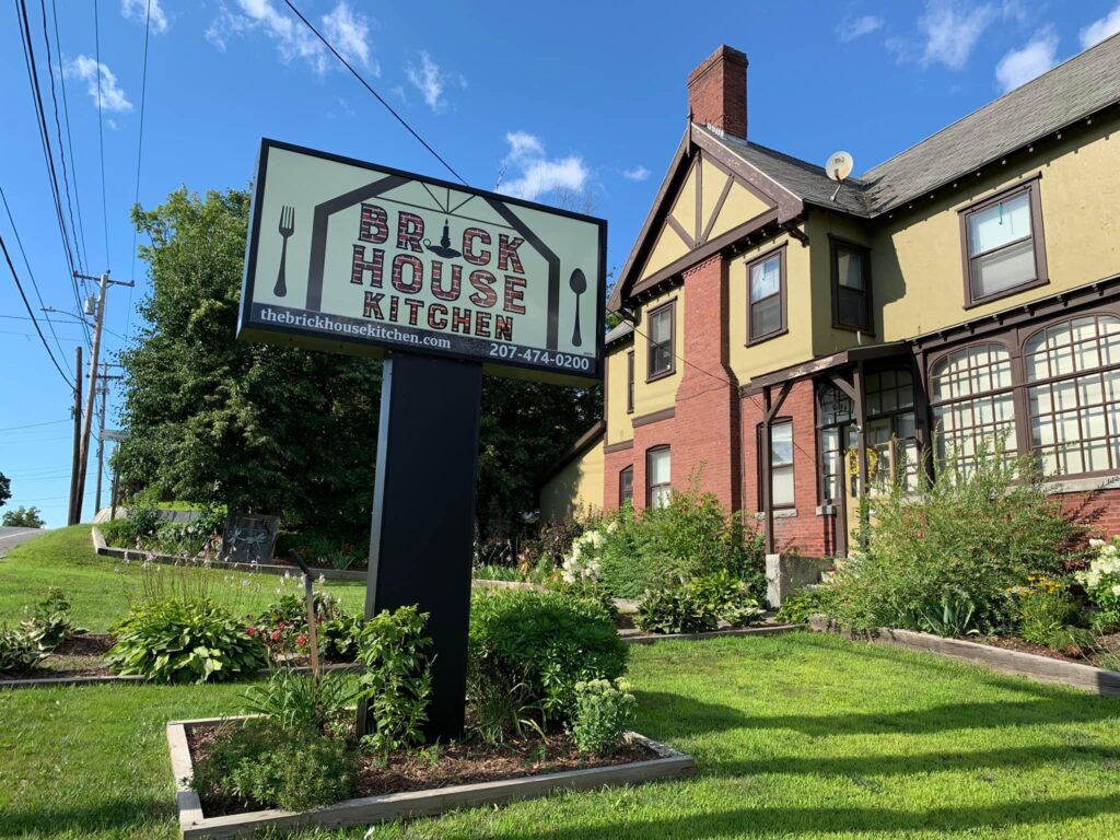 Outside of Brick House Kitchen in Skowhegan, brick building and business sign with lush greenery on a bright sunny day