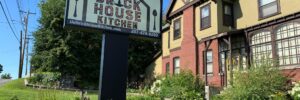 Outside of Brick House Kitchen in Skowhegan, brick building and business sign with lush greenery on a bright sunny day